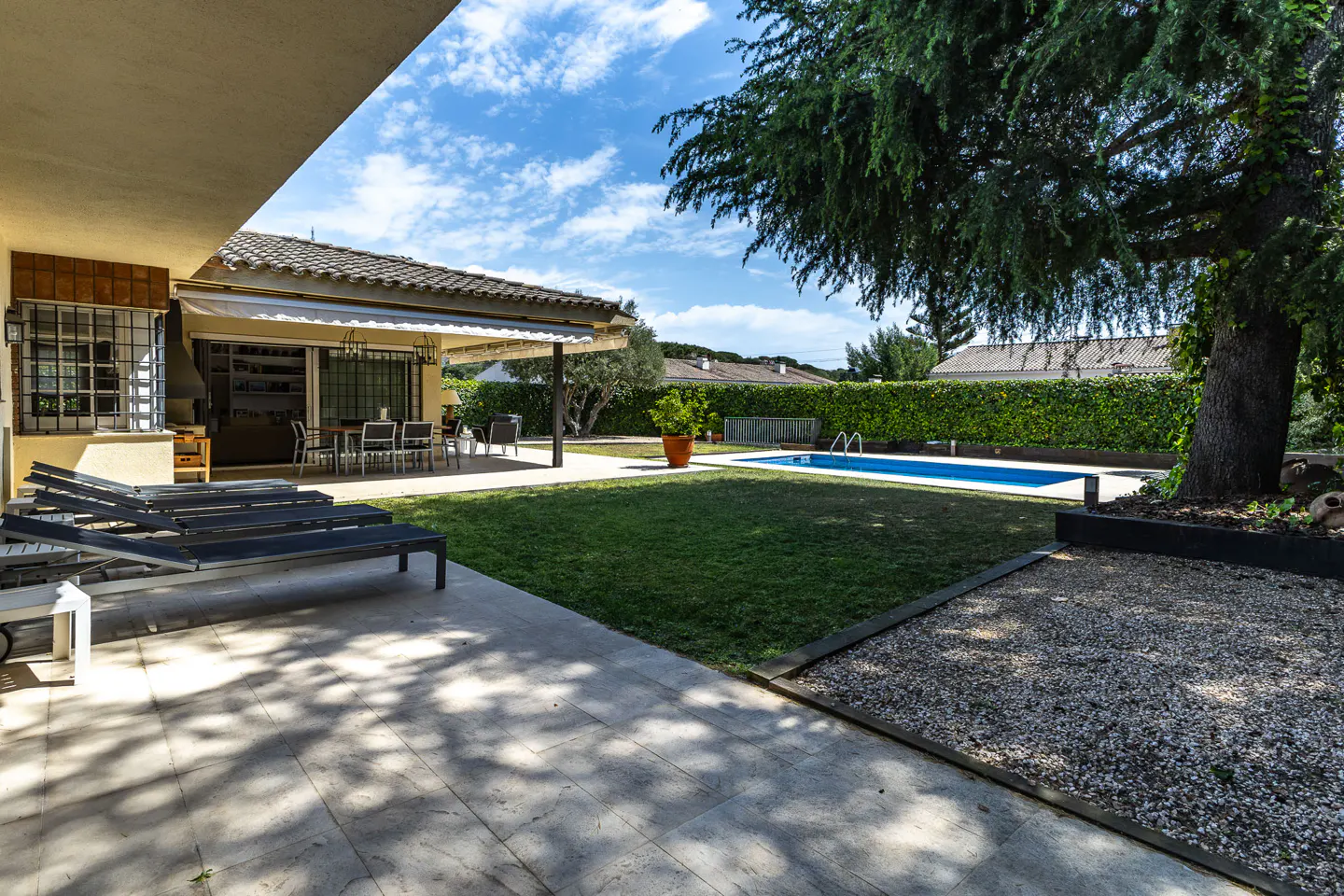 Backyard patio with lounge chairs, dining table, green lawn, and blue pool, all under a partly cloudy sky.