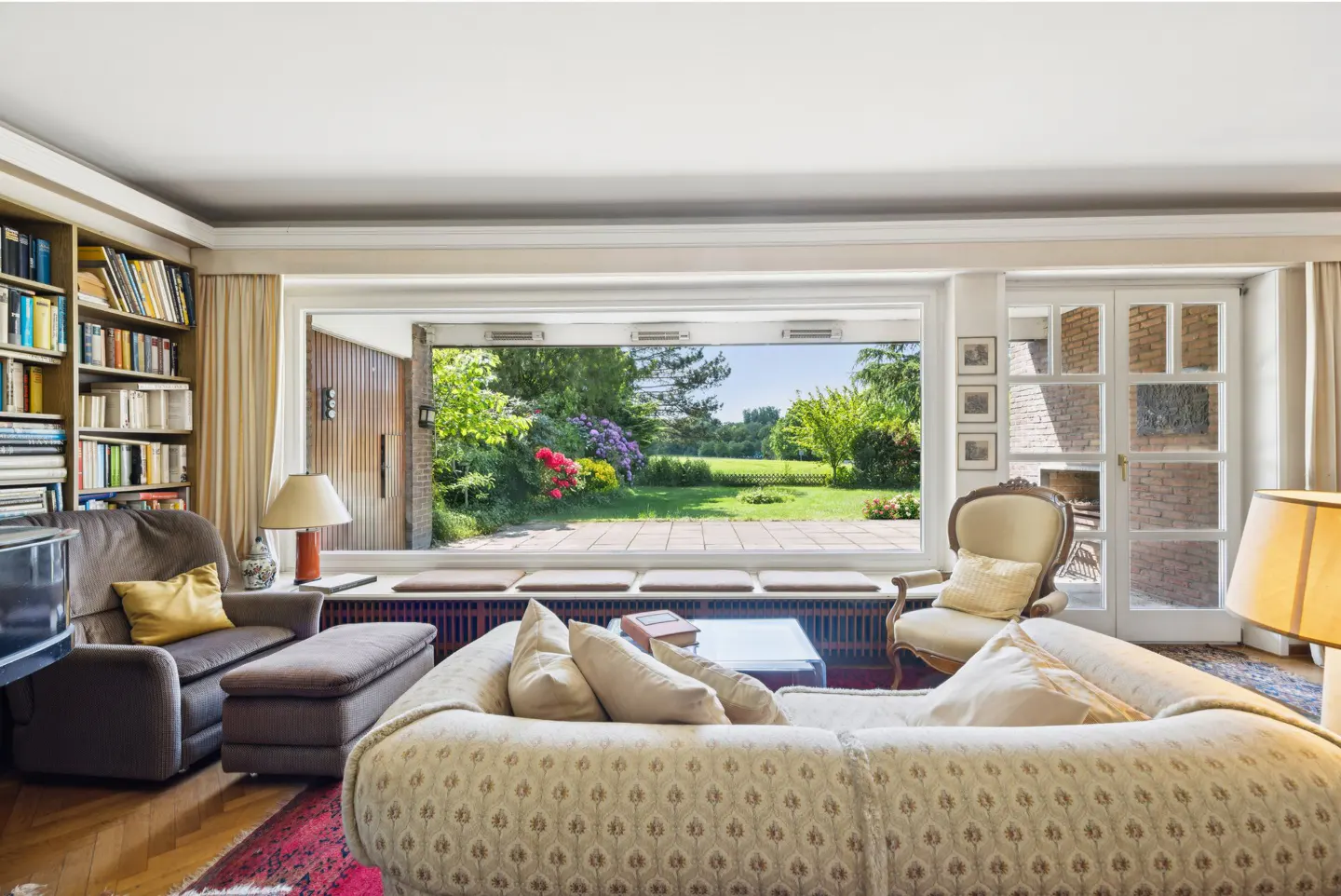 Living room with a large window view of a garden. A sofa, chair, and bookcase are visible.
