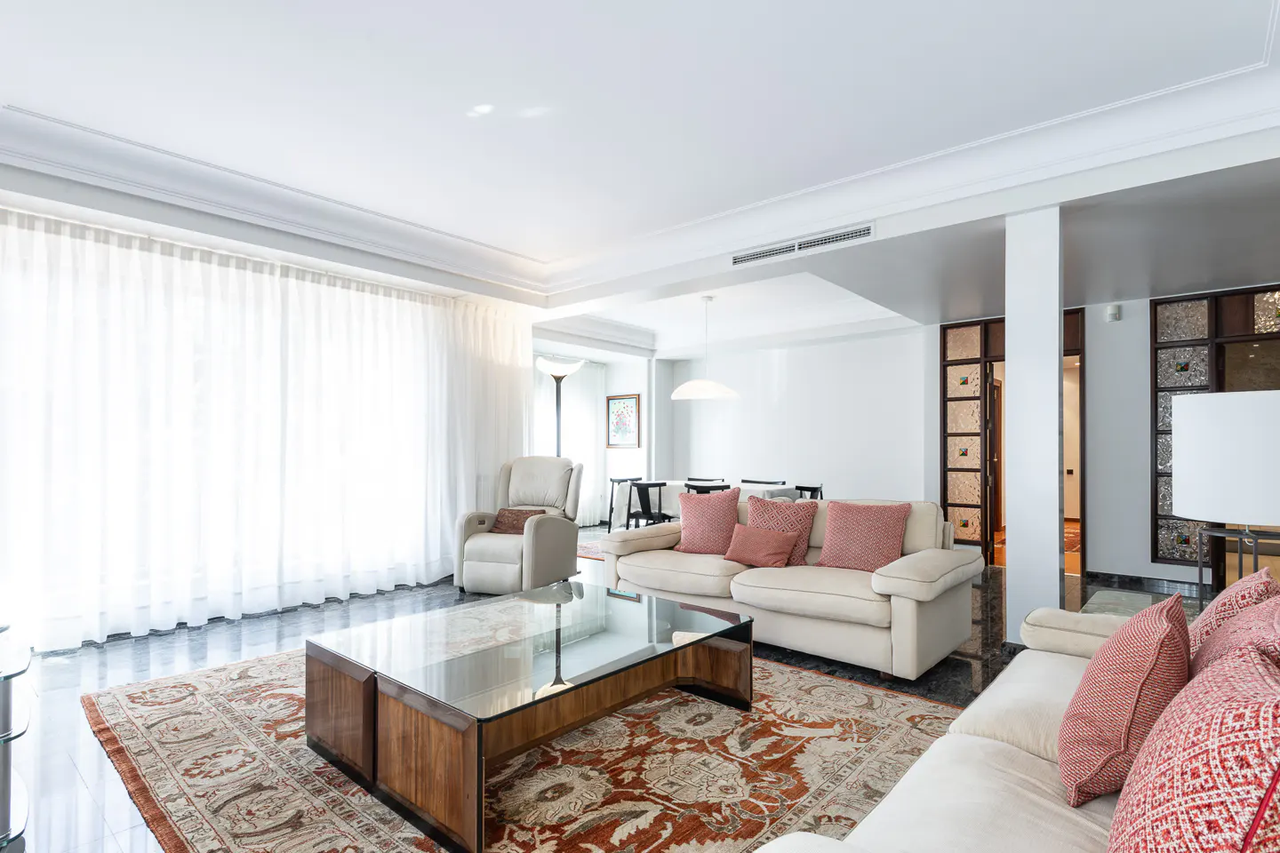 Bright living room with white sofas, red patterned pillows, and a glass-topped wood coffee table on an ornate rug. White curtains cover the windows.