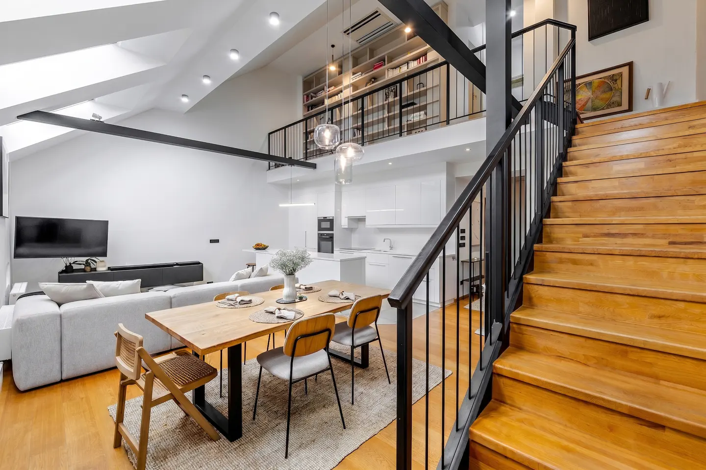 Bright, modern interior with wooden stairs, black railings, and a dining table with chairs. A white kitchen and living area are visible in the background.