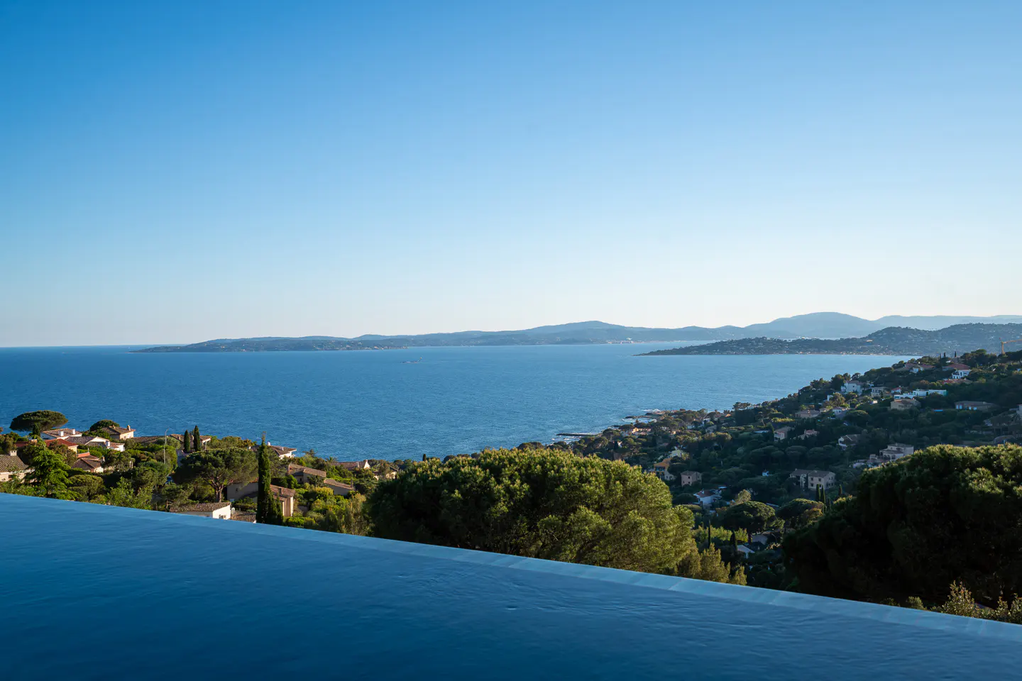 Scenic view of the French Riviera from an infinity pool, with blue water, green trees, and houses dotting the hillside.