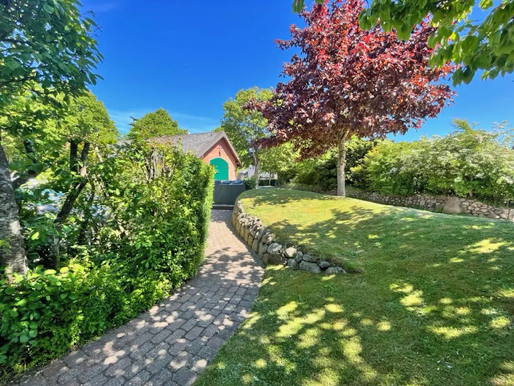 A stone path leads through a lush garden toward a small brick building with a green door under a bright blue sky.