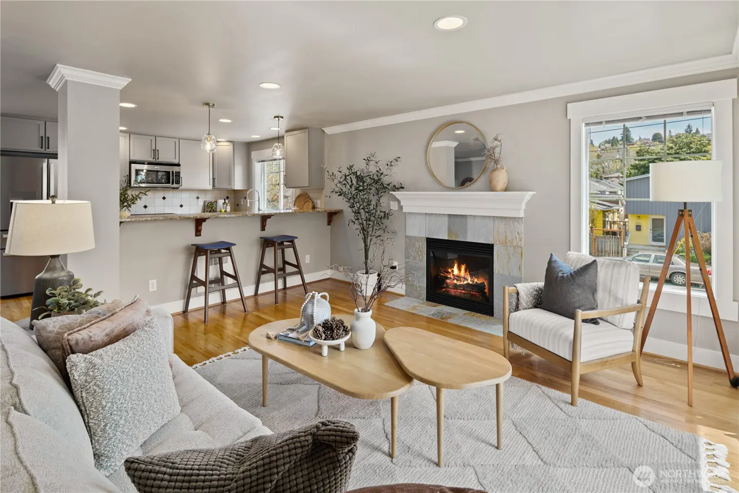 A bright living room with a fireplace, a gray sofa, a wooden coffee table, and a kitchen in the background.