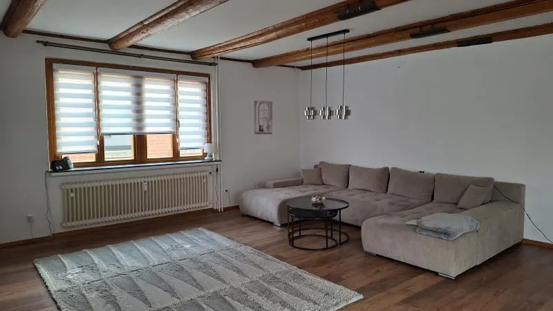 Living room with a beige sectional sofa, round coffee table, and gray rug on a wood floor. The ceiling has exposed wooden beams and modern pendant lights. A window with blinds is on the left.