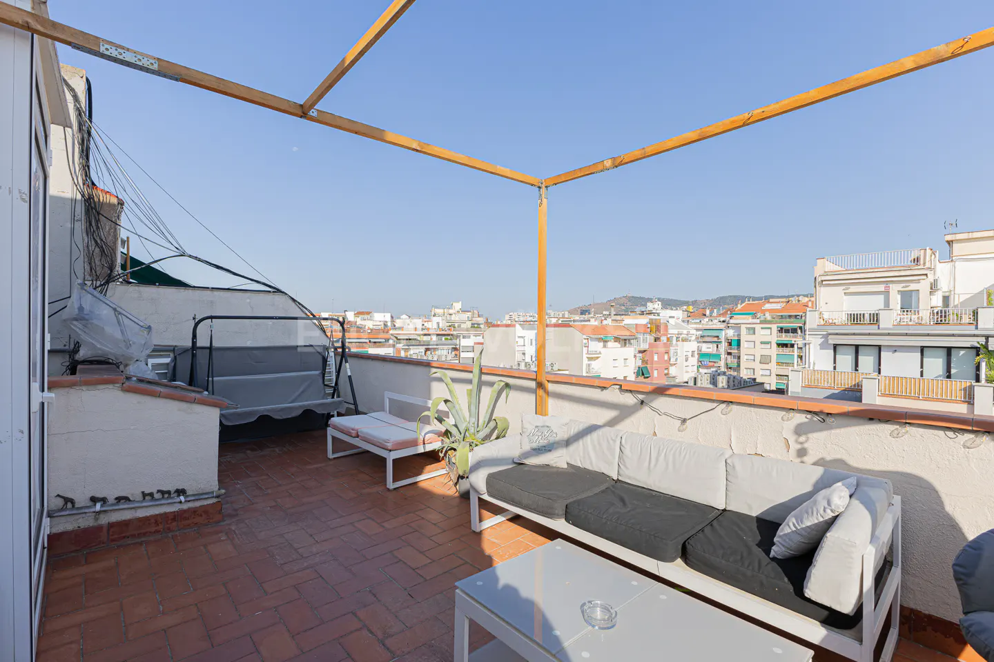 Rooftop patio with brick flooring, white sofa, lounge chair, and table. City buildings are visible in the background under a blue sky.