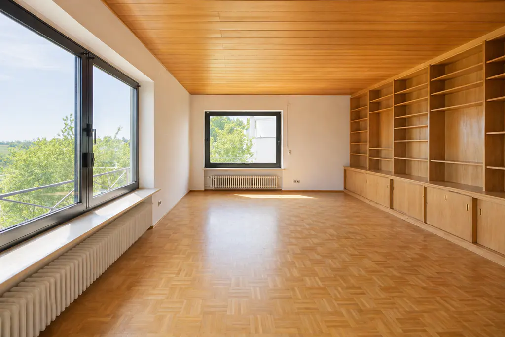 Bright, empty room with parquet floors, wood paneled ceiling, and built-in shelves. Large windows offer a view of greenery.