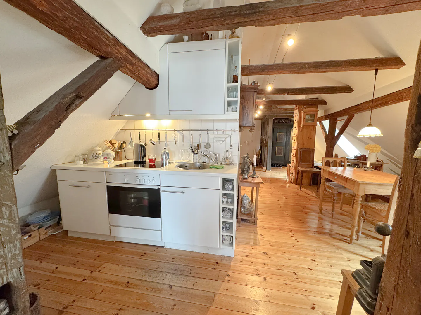 Attic apartment with wooden beams, light wood floors, and white kitchen. Dining table and chairs in the background.