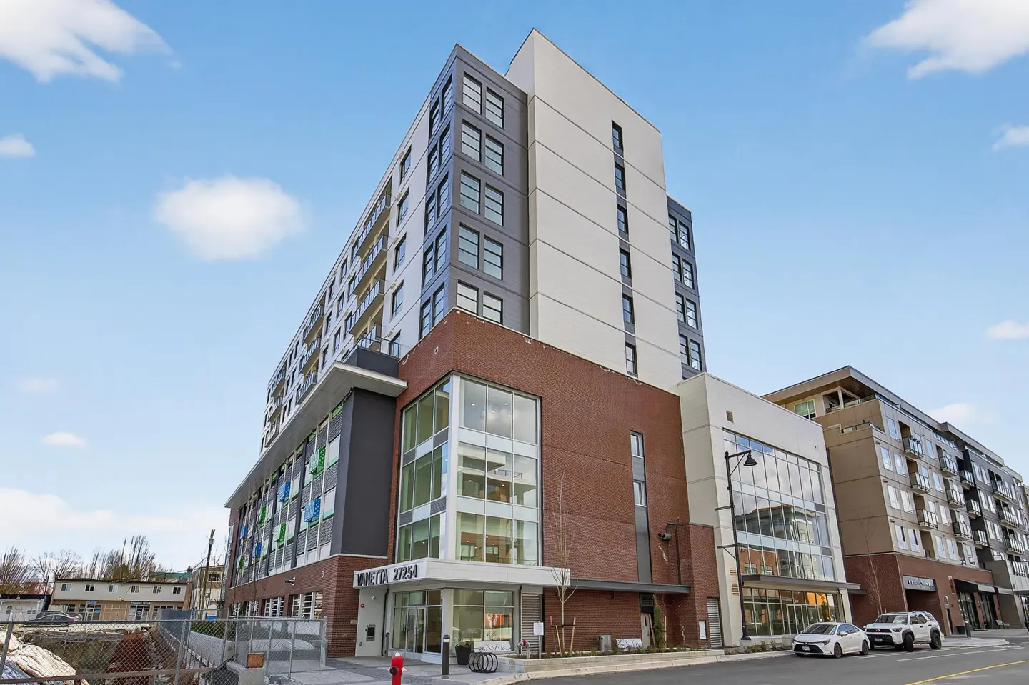 Modern multi-story apartment building with red brick and white facade, glass windows, and street-level parking.