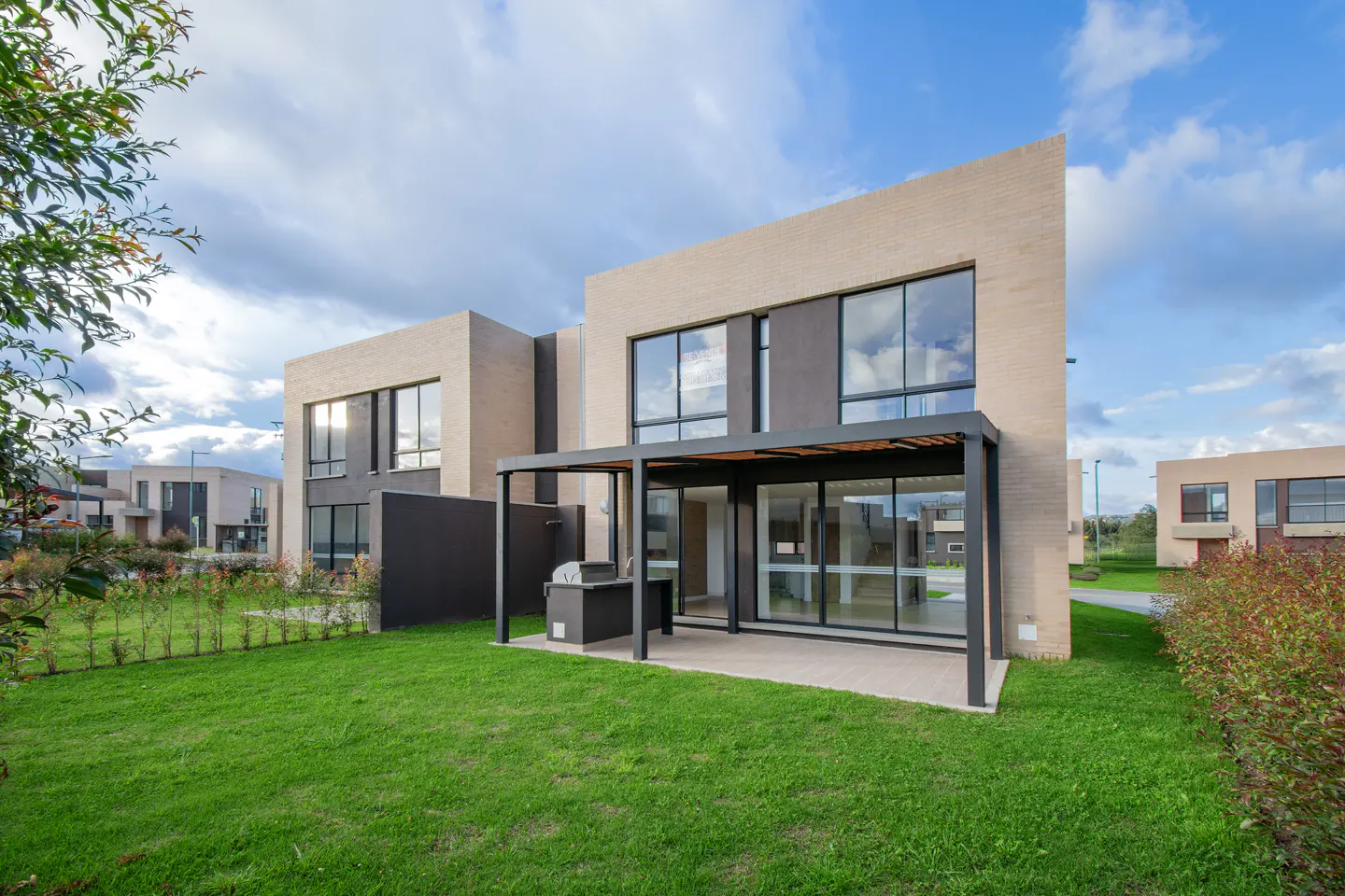 Modern two-story home with beige brick and dark gray accents, large windows, and a patio with a grill. Green lawn and cloudy sky.
