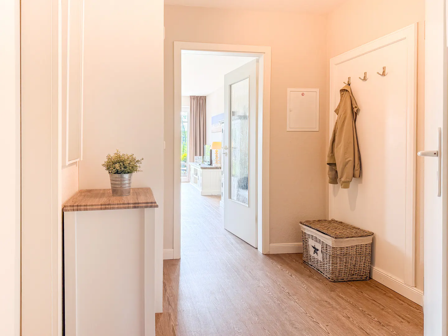 Bright hallway with wood floors, white walls, and an open door leading to a living room. A coat hangs on hooks near a wicker basket.