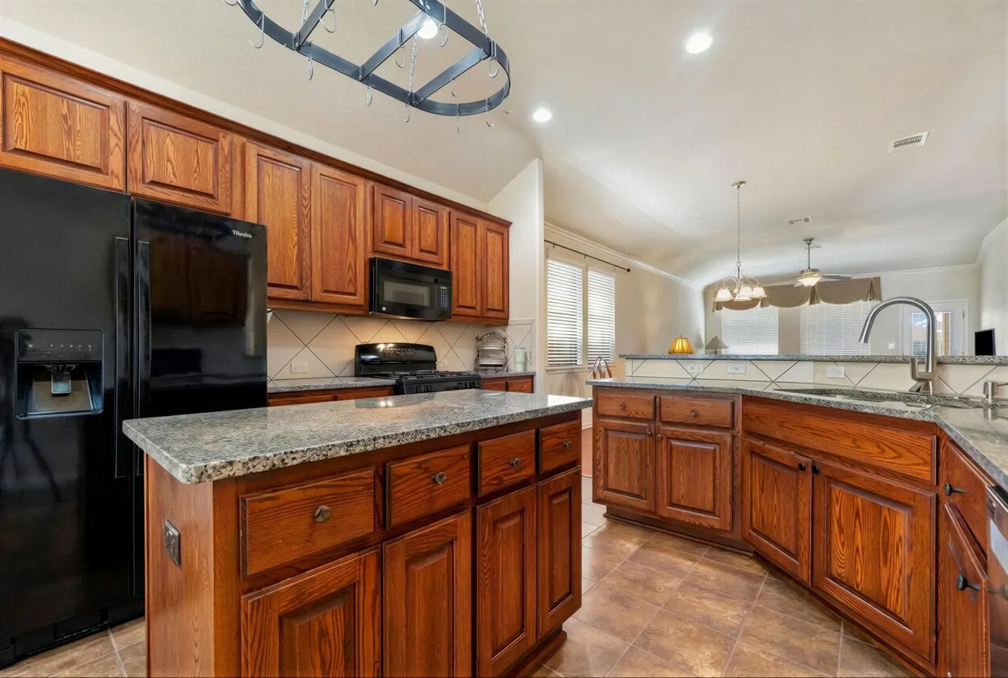 A kitchen with wood cabinets, granite countertops, and a black refrigerator. A metal pot rack hangs above an island.