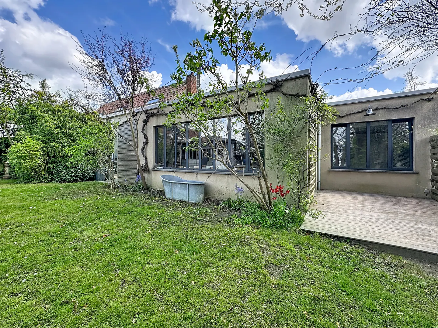 A one-story house with a green lawn, trees, and a blue sky with white clouds. A metal tub sits outside the window.