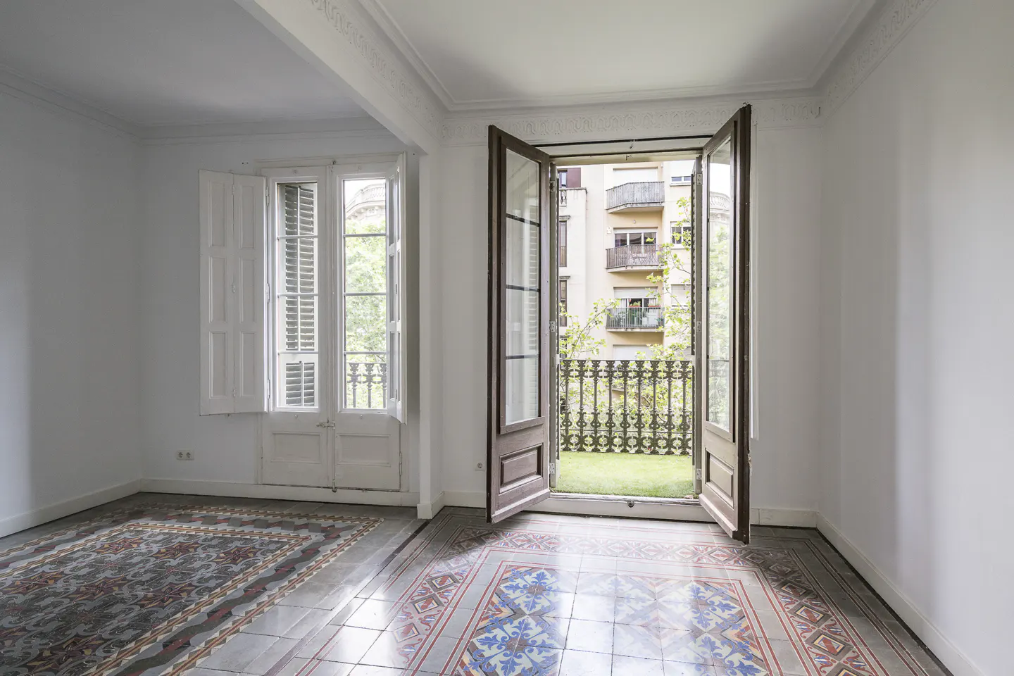 Empty room with white walls, patterned tile floors, and open French doors leading to a balcony overlooking city buildings.