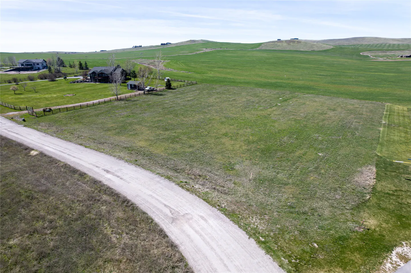 Aerial view of a grassy lot with a dirt road, green fields, and a few houses in the background under a blue sky.