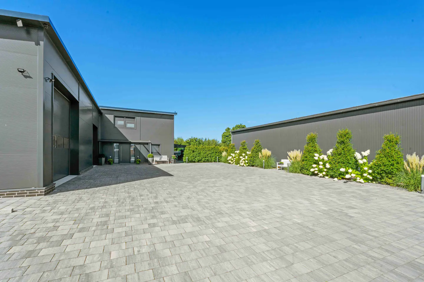 Exterior view of a gray industrial building with a paved courtyard under a clear blue sky.