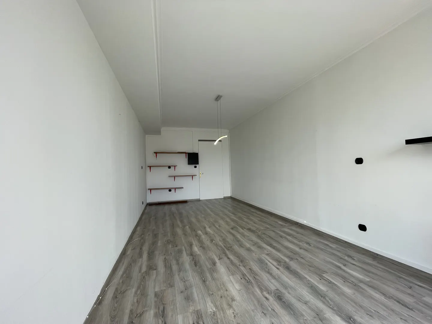 An empty room with gray wood floors, white walls, and a modern light fixture. There are red shelves on the left wall.