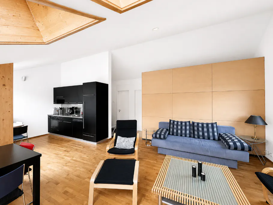 Bright apartment interior with wood floors, black kitchen cabinets, and a blue sofa with plaid pillows. A wooden coffee table sits in front.