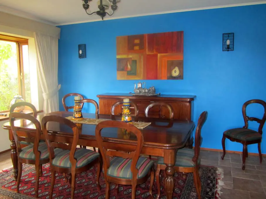 A dining room with a blue wall, a wooden table with chairs, and a red patterned rug. An abstract painting hangs above a wooden sideboard.