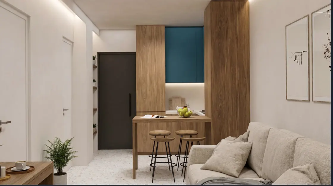 A modern apartment interior with a beige sofa, wooden kitchen cabinets, and a small dining table with two stools. Two framed art pieces hang on the white wall.