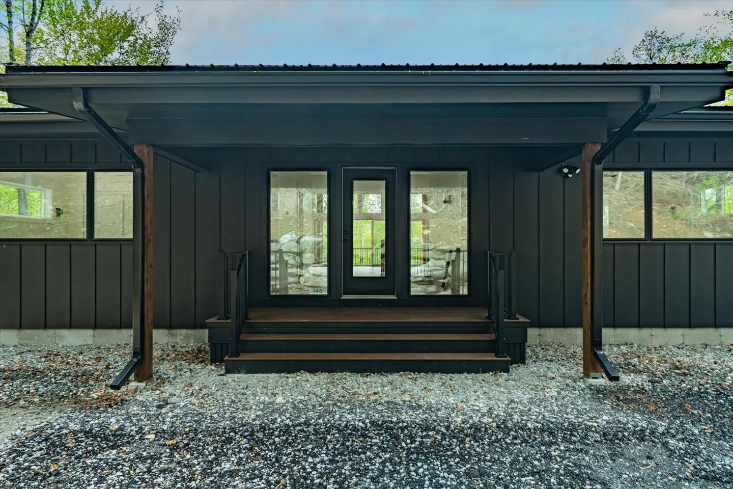 Modern dark brown house with a black door and windows, and wooden steps leading to the entrance. The ground is covered in gravel.