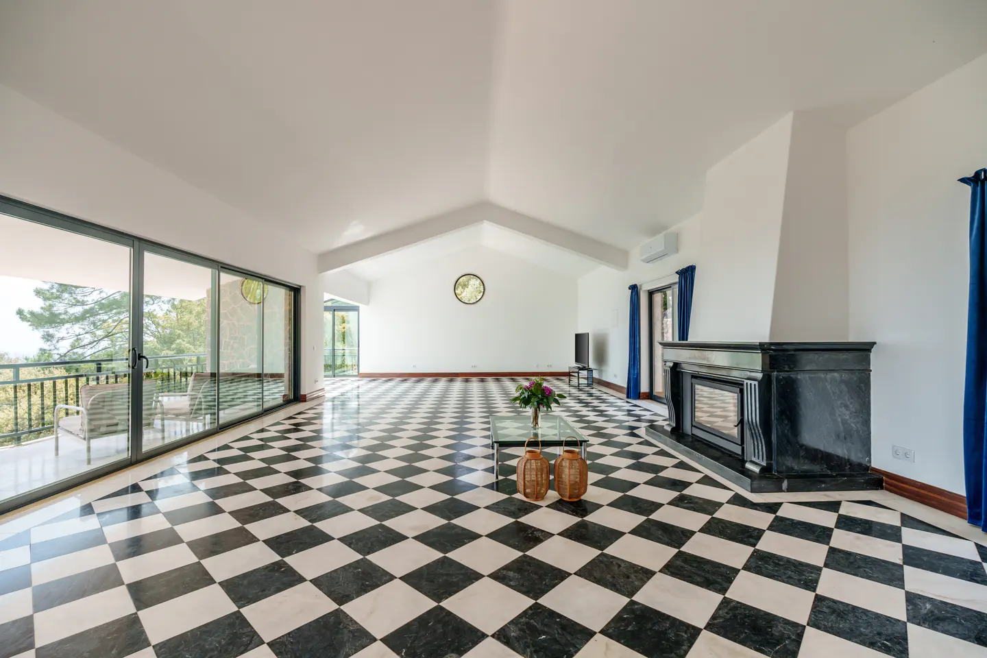 Bright living room with black and white checkered floor, fireplace, glass table, and large windows to a balcony with green trees.