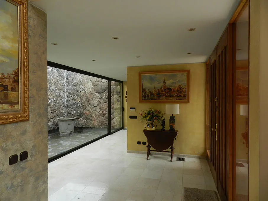 Hallway with white tile floor, yellow walls, and a stone courtyard visible through a large window. Paintings and a table with flowers decorate the space.