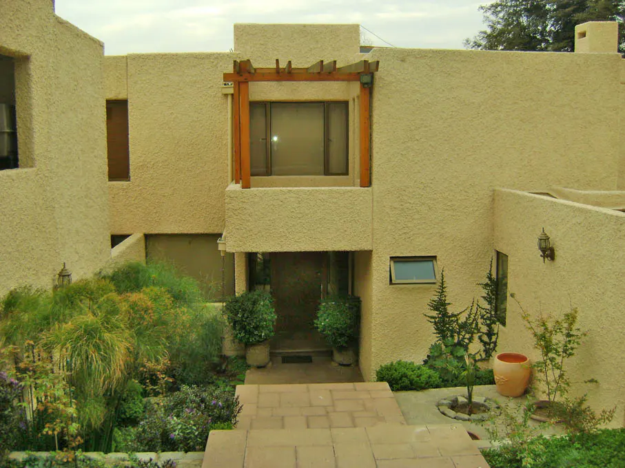 Beige stucco house with a wooden pergola over the front window, and stone steps leading to the front door. Lush green landscaping.