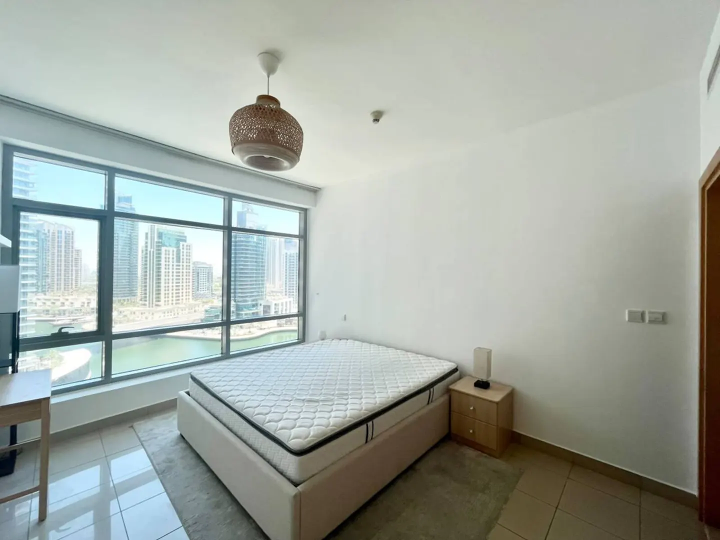 Modern bedroom with a mattress on a bed frame, a wooden nightstand, and floor-to-ceiling windows overlooking city towers.