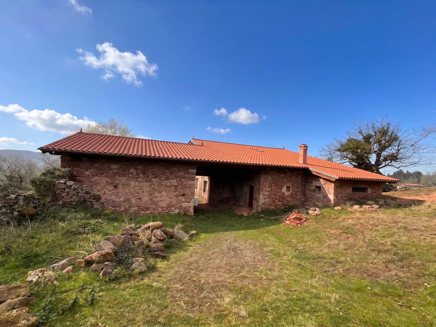 Exterior view of a one-story stone house with a red tile roof under a blue sky with scattered clouds.