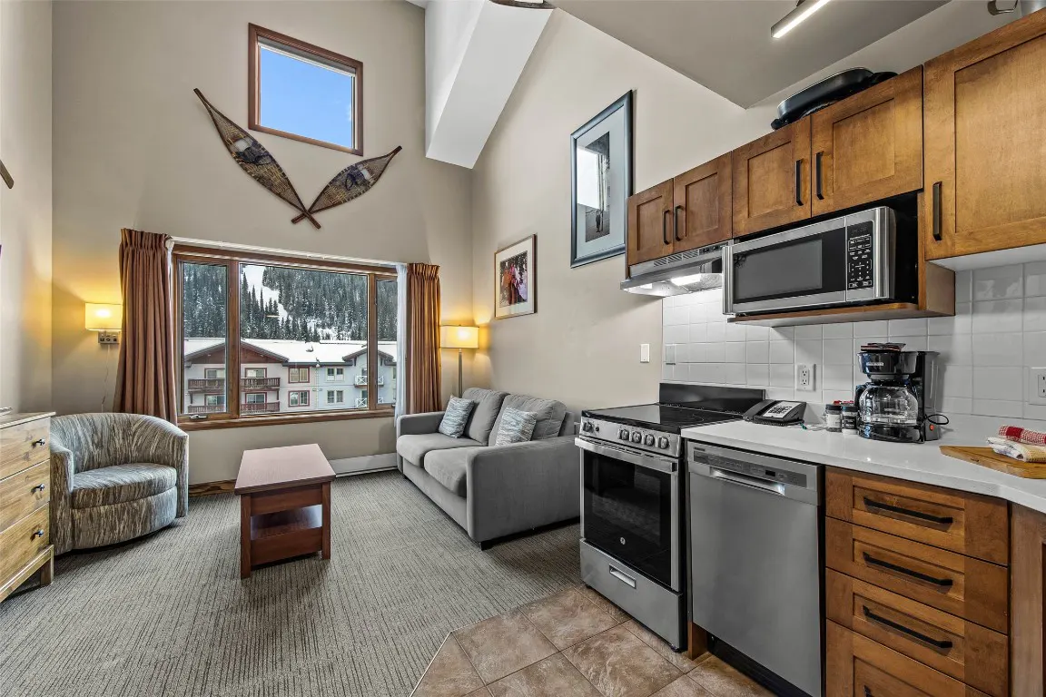 Interior view of a condo with a living room and kitchen. A window shows a snowy mountain view. Snowshoes hang on the wall.