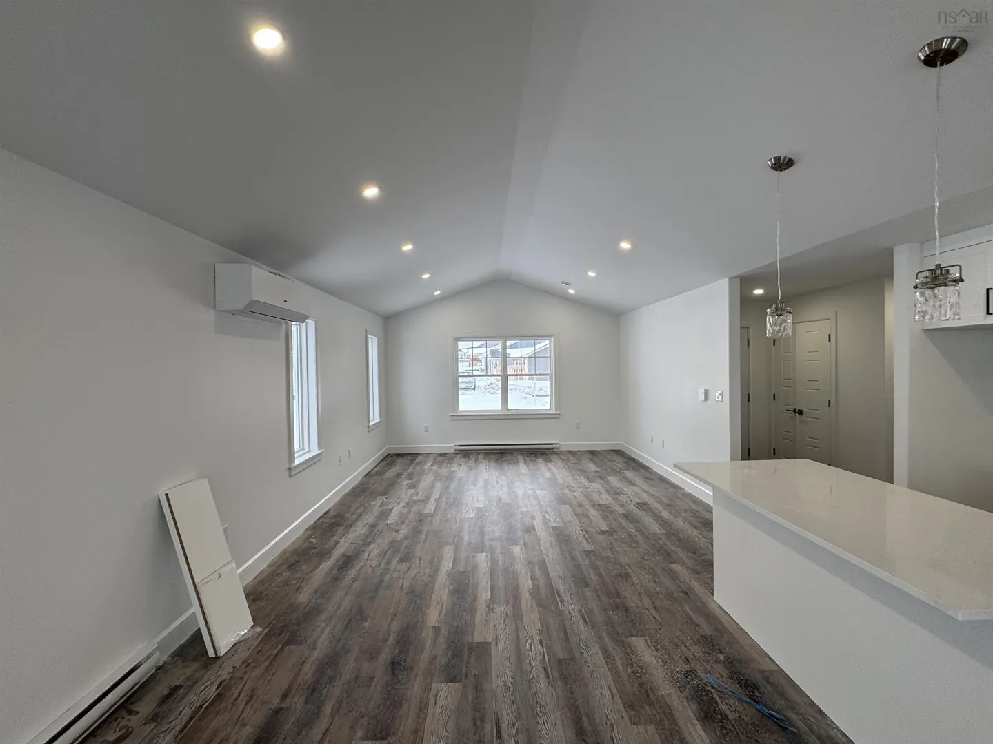 Bright, open-concept living space with gray wood floors and white walls. Recessed lighting, windows, and a kitchen island are visible.