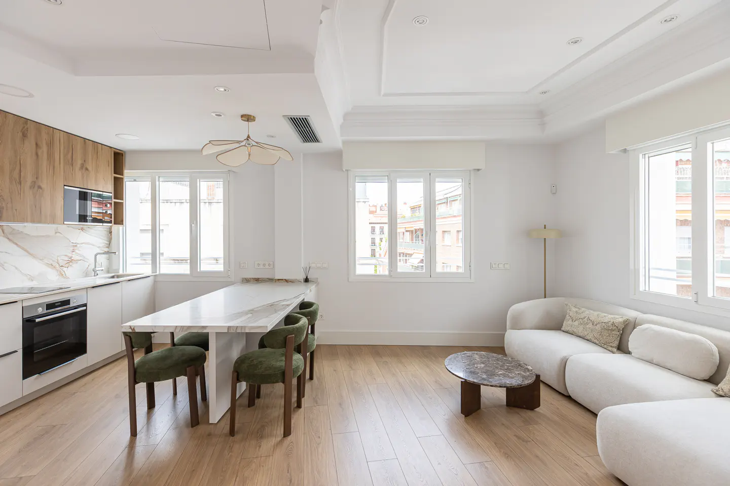 Bright, modern apartment with white walls, wood floors, and large windows. Kitchen with marble counters, green chairs, and a white sofa.