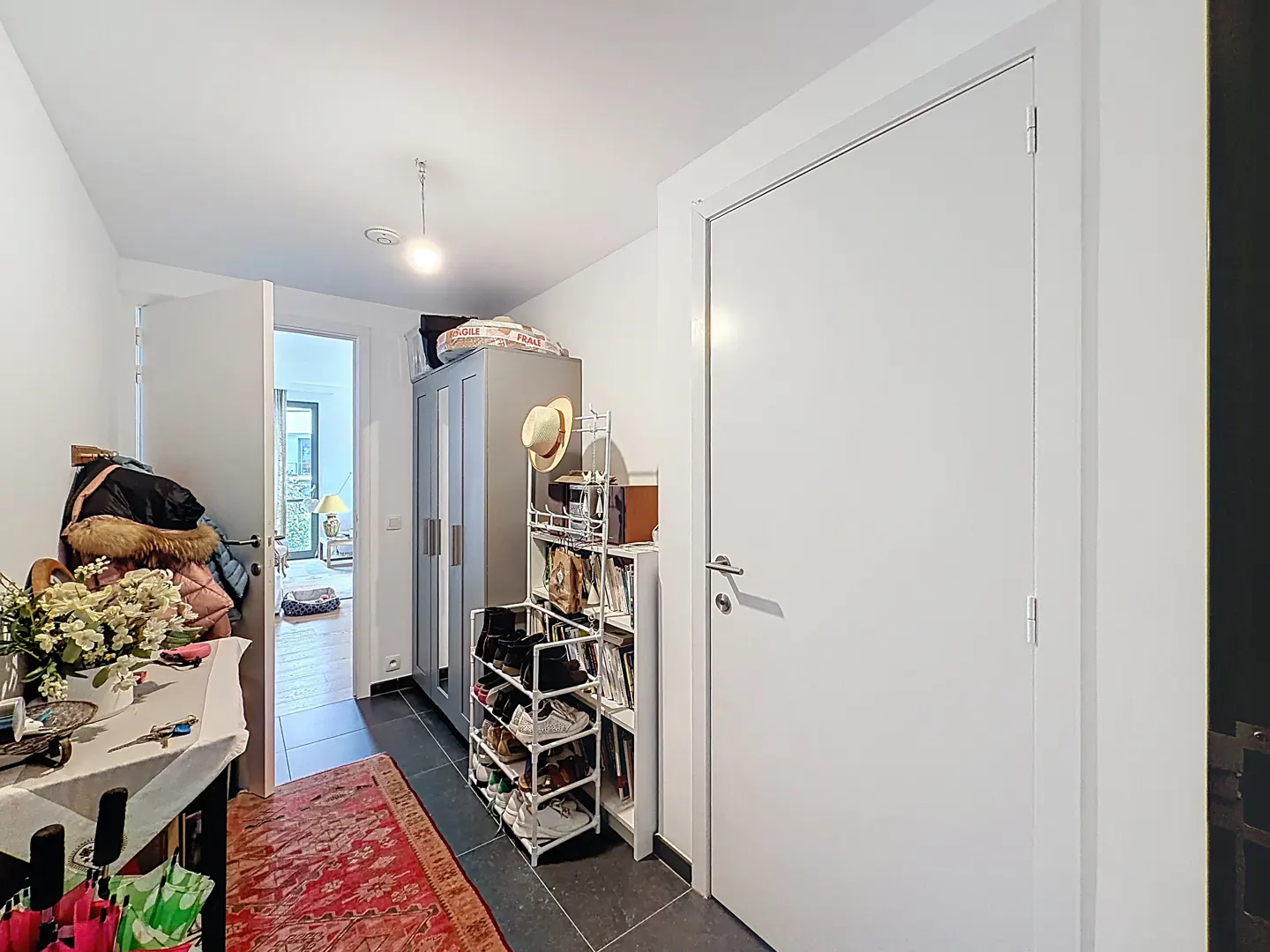 A bright hallway with a white door, grey wardrobe, and shoe rack. A red rug lies on the dark tile floor.