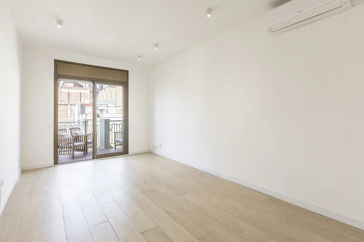 Bright, empty room with white walls, light wood floors, and sliding glass doors to a balcony with outdoor furniture.