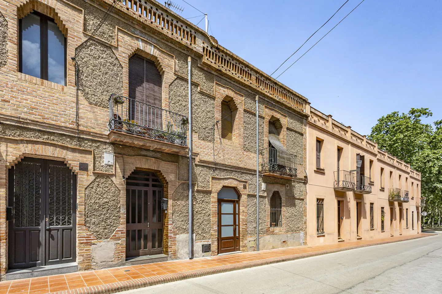 Row of brick and stucco buildings with arched windows and doors, some with small balconies, along a street.