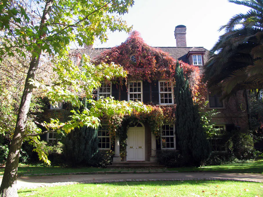 A two-story brick house with white trim is covered in green and red ivy. The house has a chimney and a white double door.