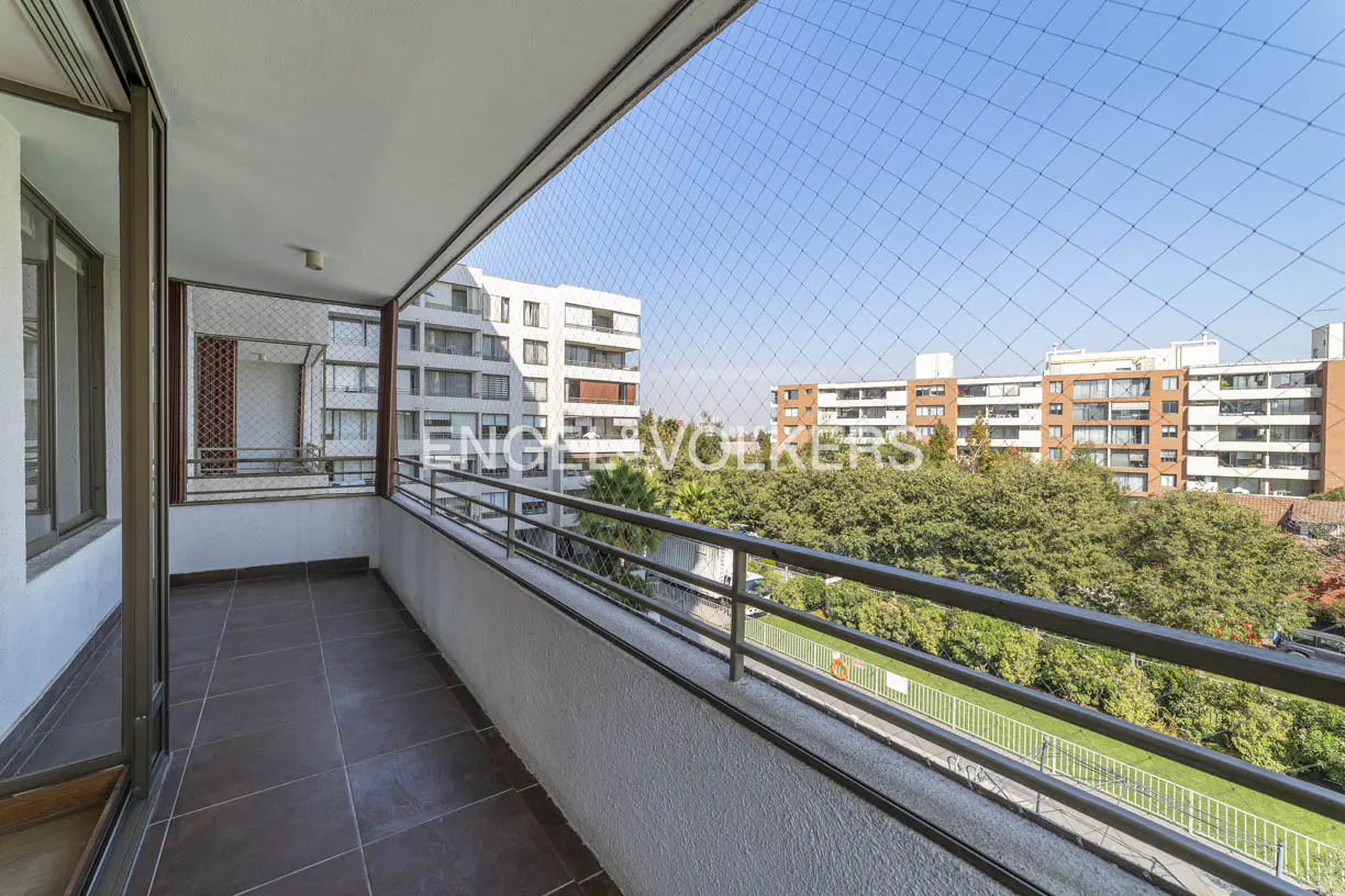Balcony view with brown tile floor, white railing, and safety net. Buildings and trees are visible in the background under a blue sky.