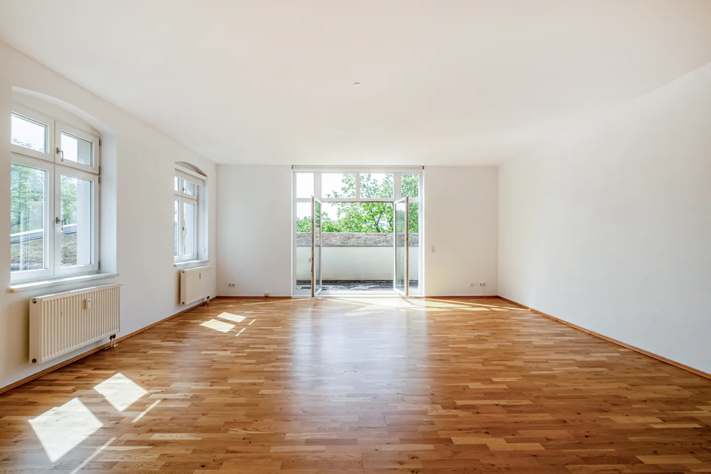Bright, empty room with hardwood floors, white walls, and two windows. Balcony doors are open, revealing greenery outside. Radiators are under the windows.