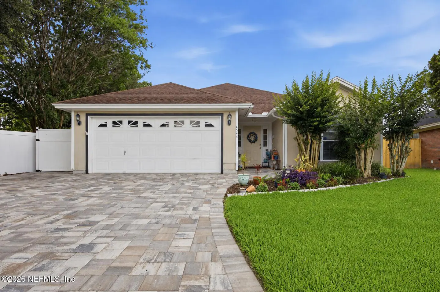 Beige single-story home with a brown roof, white garage door, and a paver driveway. Green lawn and blue sky.