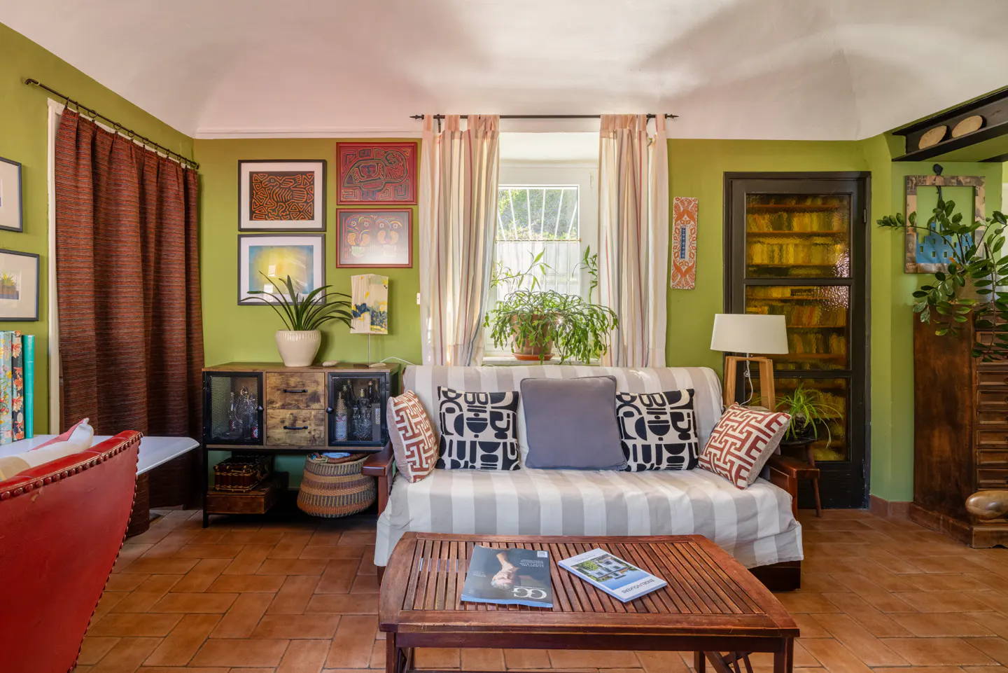 Eclectic living room with green walls, terracotta tile floor, and a striped sofa with patterned pillows. Art adorns the walls, and plants add a touch of nature.