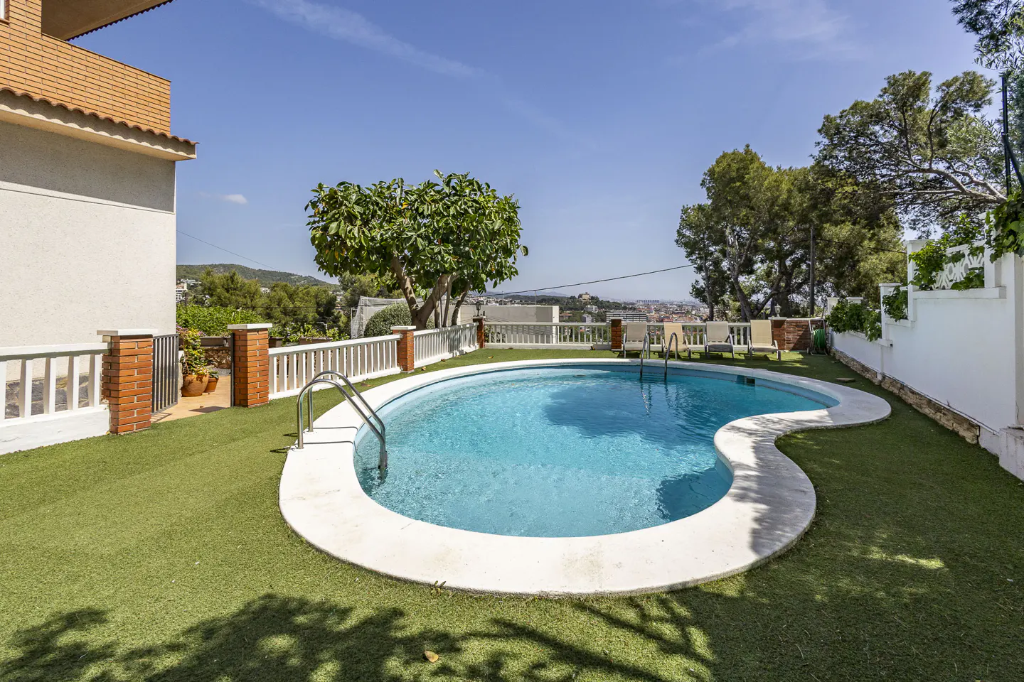 A kidney-shaped pool with turquoise water is surrounded by green turf and a white fence. Lounge chairs are visible in the background.