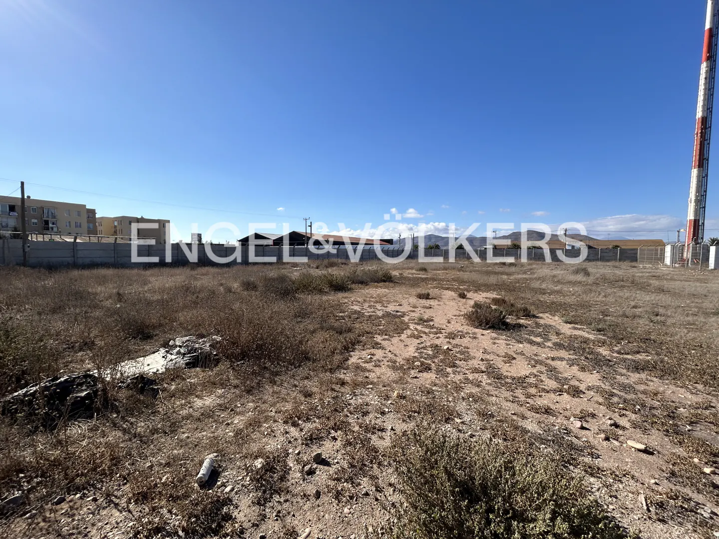 Vacant lot with dry brush under a clear blue sky. A concrete wall and buildings are in the background. A red and white tower is on the right.