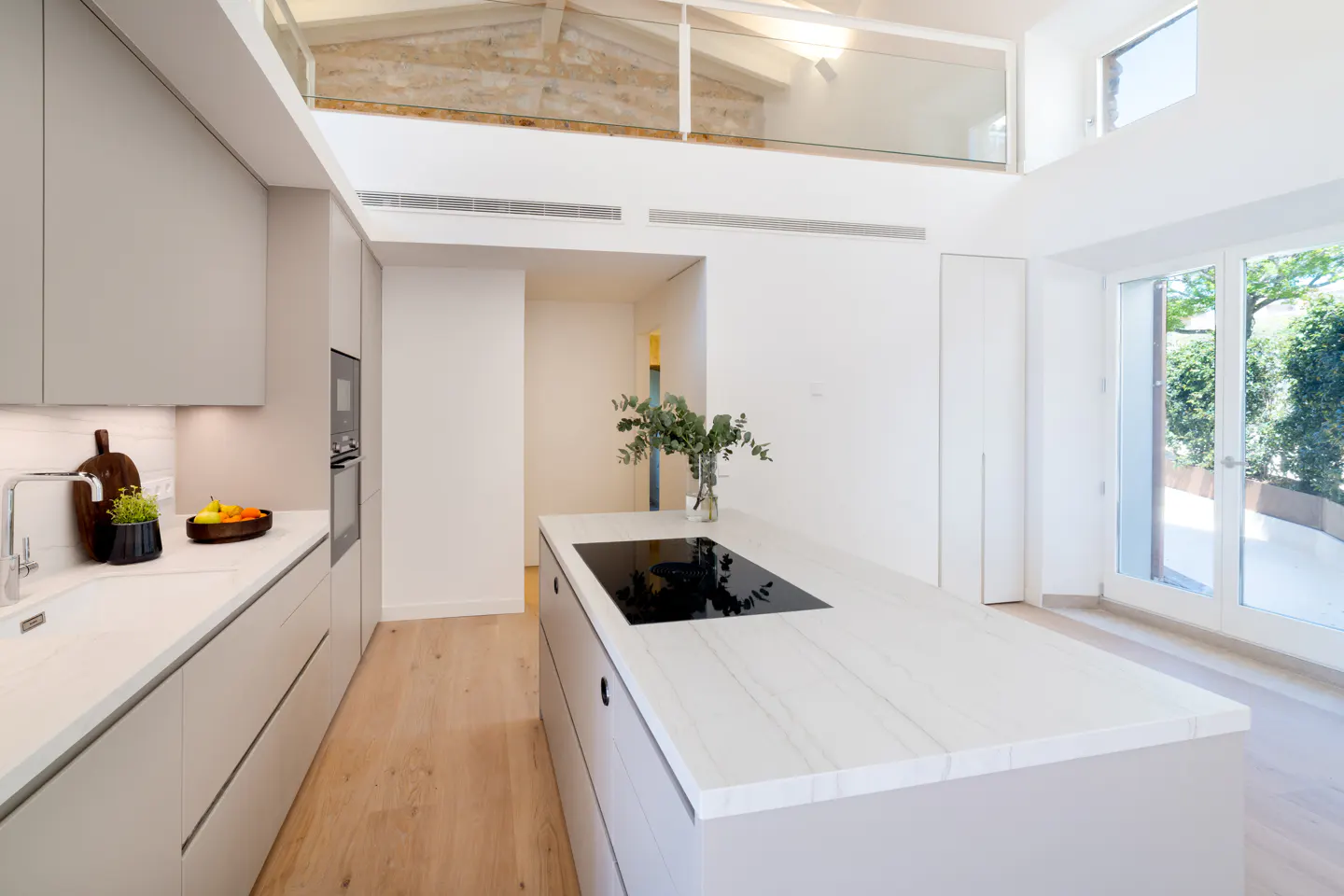 Bright, modern kitchen with white walls, light wood floors, and gray cabinets. A large island features a black cooktop and marble countertop. Glass doors lead to a green outdoor space.