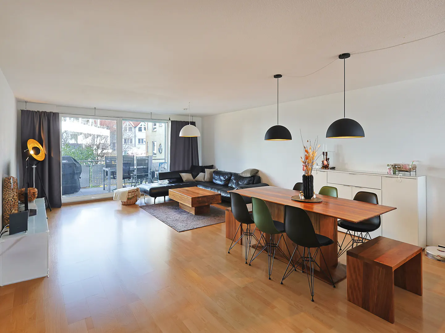 Bright, open-concept living and dining area with wood floors. Black leather sofa, wood table with chairs, and balcony with outdoor furniture.