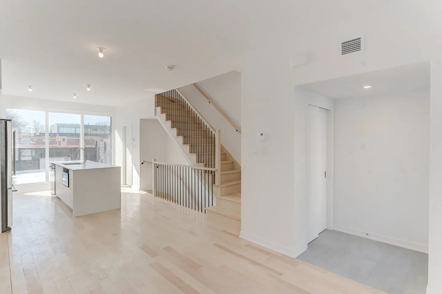 Bright, open-concept home interior with white walls, light wood floors, kitchen island, and staircase with black railings.