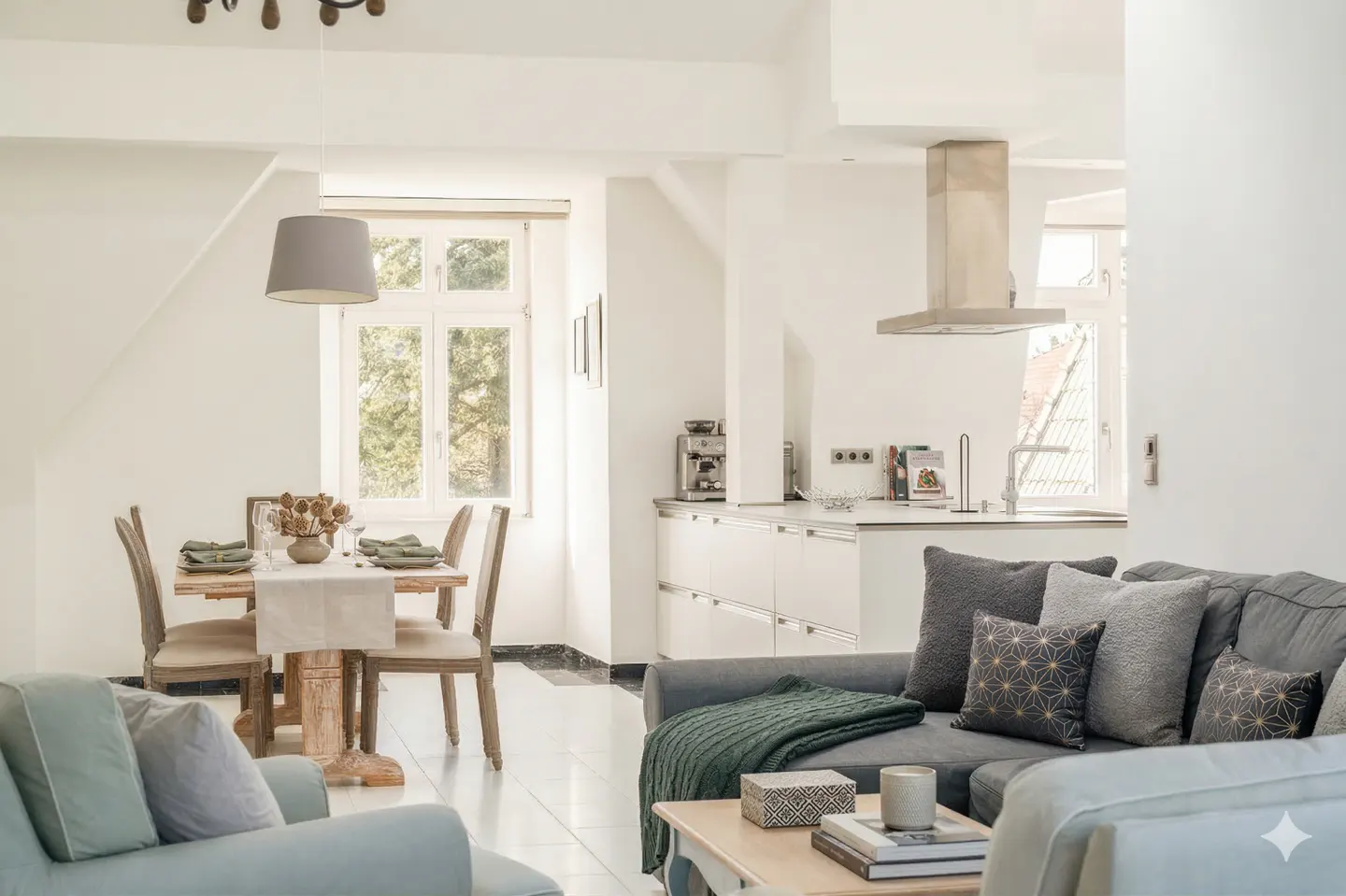 Bright, modern living space with a dining table set for four, a gray sofa with patterned pillows, and a white kitchen in the background.
