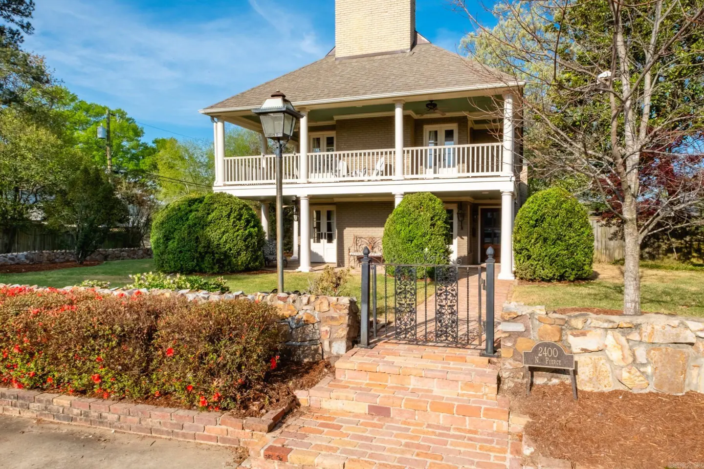 Two-story house with a porch, brick steps, and a black iron gate. The house is surrounded by green bushes and red flowers.