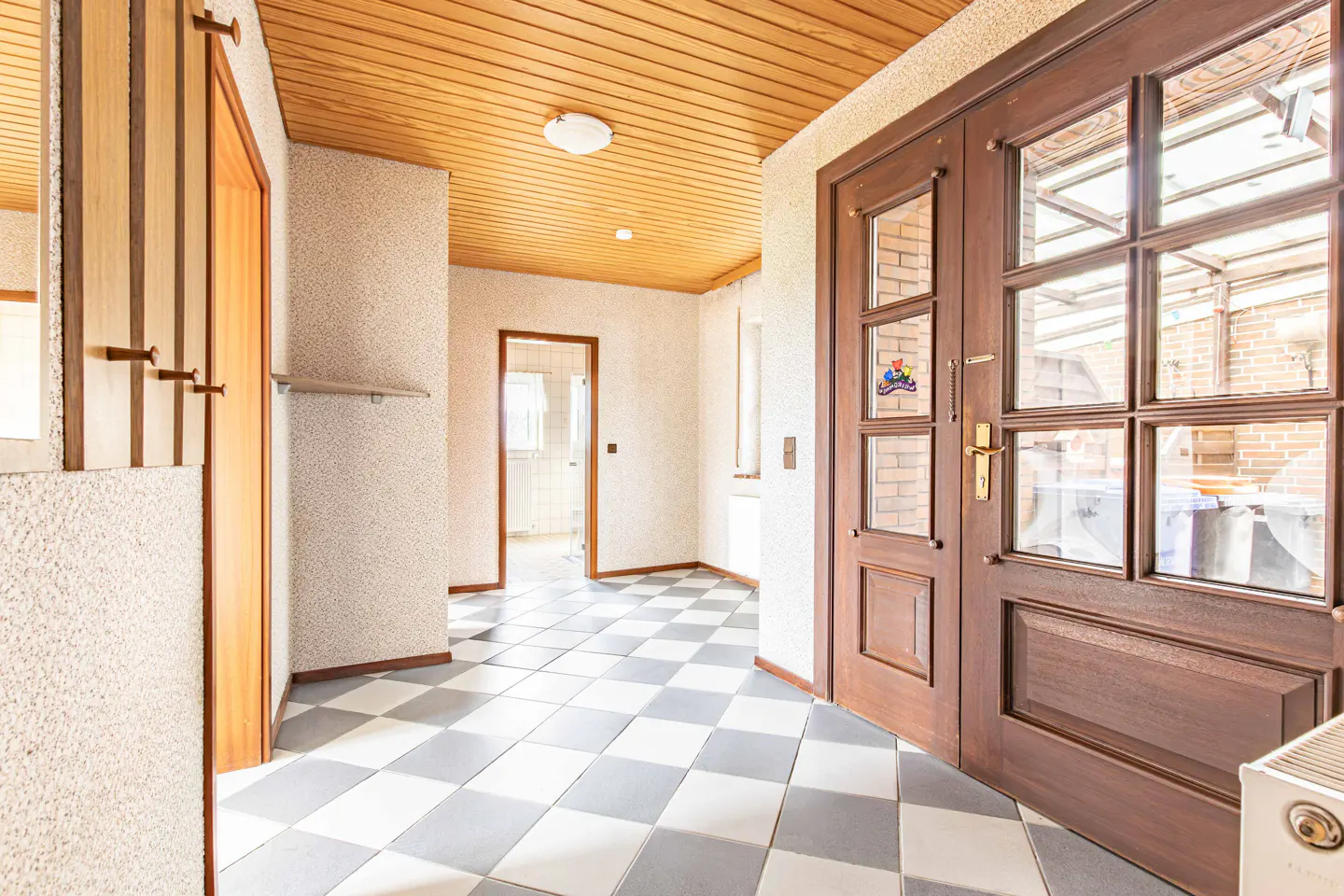 Hallway with checkered tile floor, textured walls, and wood-paneled ceiling. A glass-paneled door leads outside.