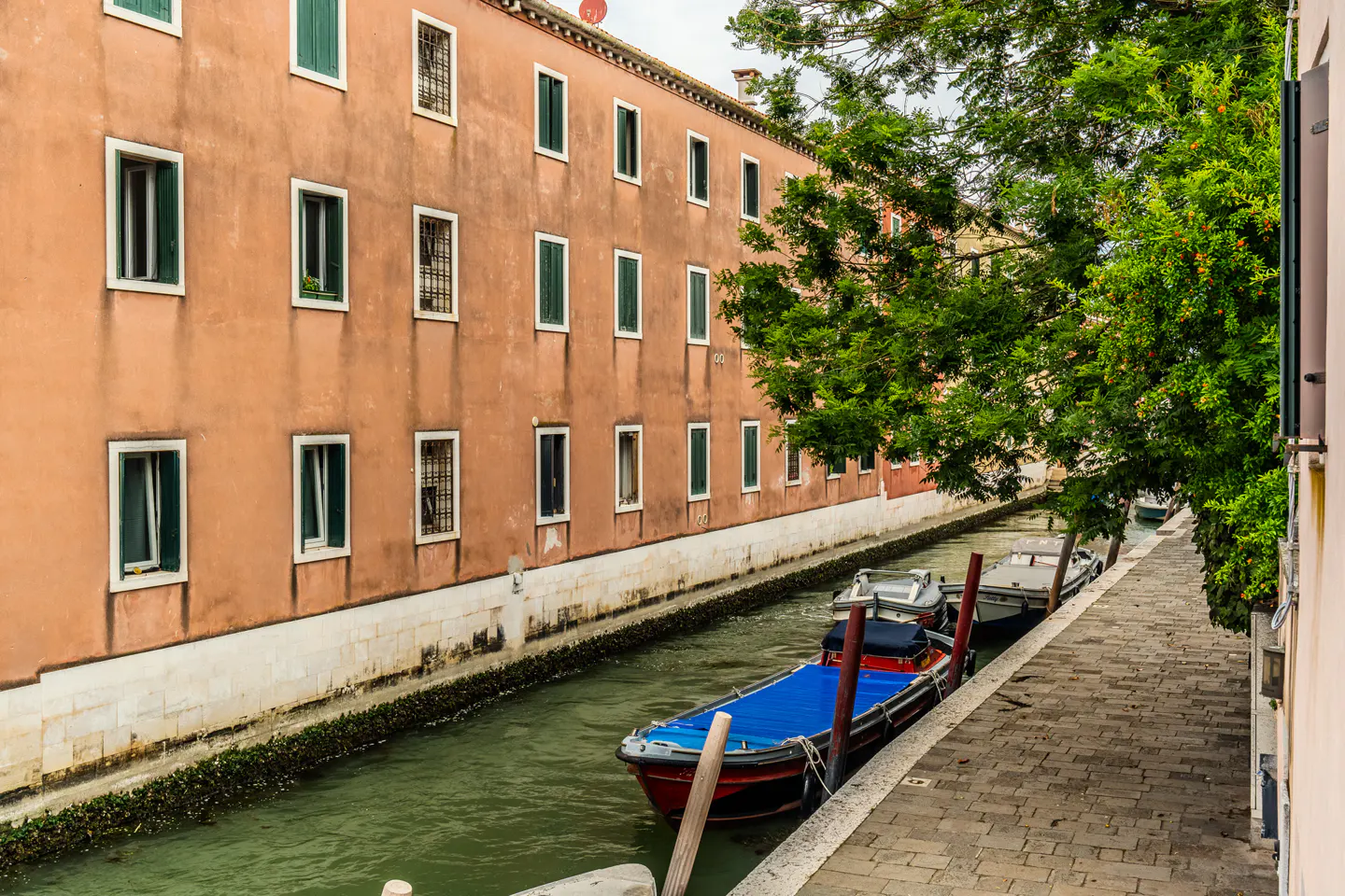 A canal in Venice with boats moored along a stone walkway next to a tall, peach-colored building with green shutters.