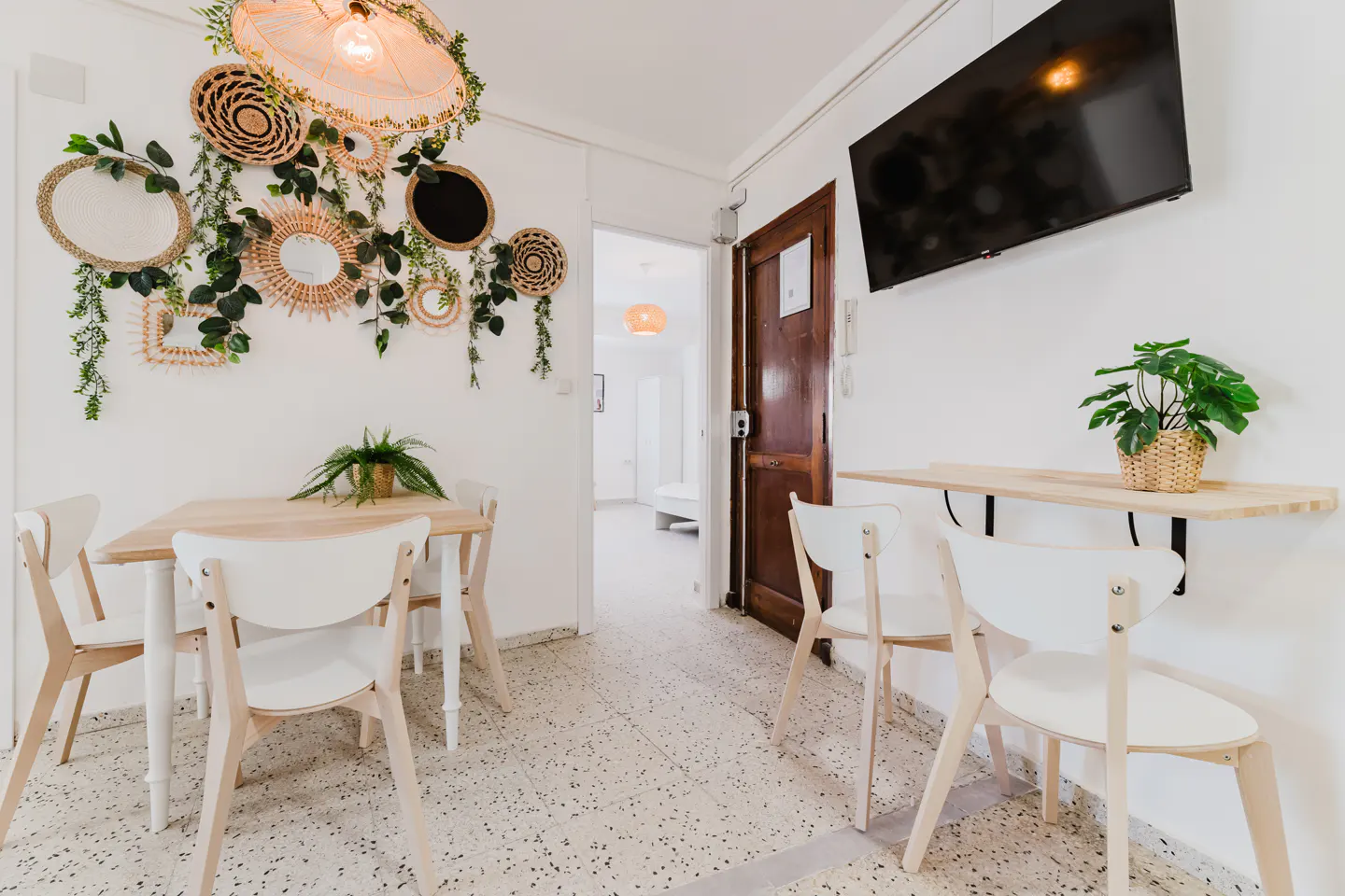 Bright dining area with white walls, terrazzo floor, and wooden furniture. Wall decor includes woven baskets, mirrors, and greenery. A TV hangs above a folding table with a potted plant.